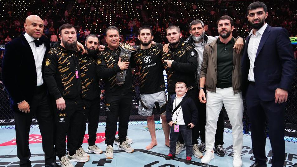 Islam Makhachev poses with his UFC title, surrounded by his team and coaches in the cage, including Khabib Nurmagomedov