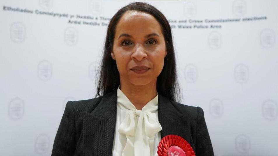 A head and shoulders picture of Emma Wools. She has black shoulder-length hair and is wearing a dark jacket with a red Labour rosette on it and a white blouse.