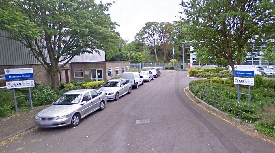 The East of England Ambulance Service Trust HQ in Melbourn. It shows a business park with low-lying buildings and with cars parked on the left. On either side of the road is a trust sign.