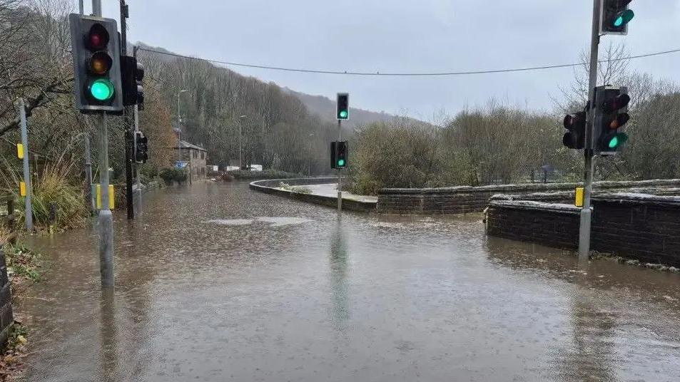 Burnley Road at Callis Bridge is shown heavily flooded as water covers the entire road. Traffic lights are shown as is a stone bridge which crosses the River Calder.