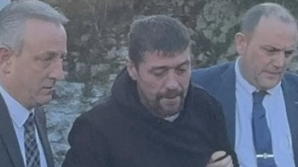 Joe Butler, being escorted by two men in suits who are holding him by the arms. He is wearing a dark coloured hooded jumper and behind him is a stone wall. 