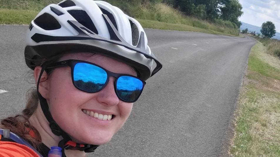 Emma Davis in a selfie, with blue sunglasses and wearing a black and white cycle helmet on an empty country road