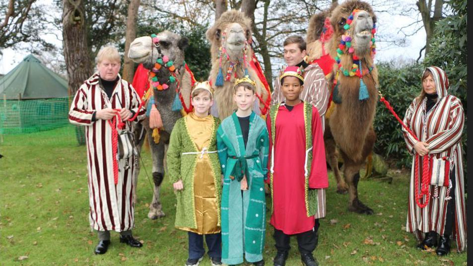 Three children aged around 11 or 12 dressed as the Three Kings from the nativity pose in front of three large camels which are flanked by their handlers. 