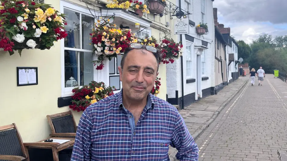 Hossey is standing on the street in Bewdley by the pub. He has a checked shirt on and glasses on his head and dark short hair. The cobboled street can be seen in the background.