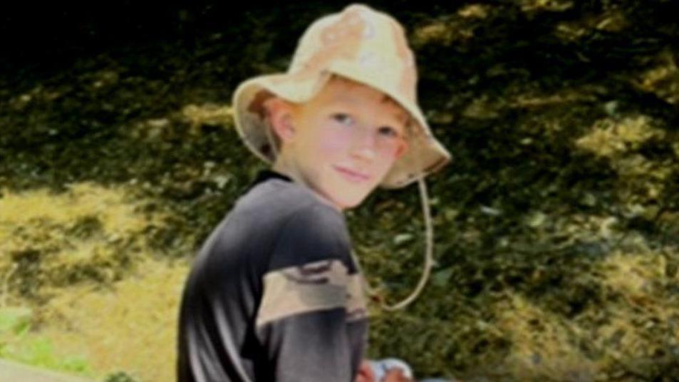 Zac Roe looking over his shoulder at the camera. He is wearing a dark top and a floppy summer hat. The background shows tree shadows on grass.