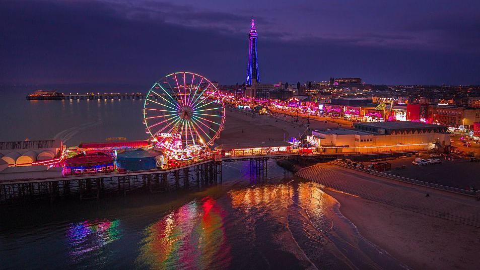 Blackpool pier is shown lit up by ferris wheel and lights along the promenade