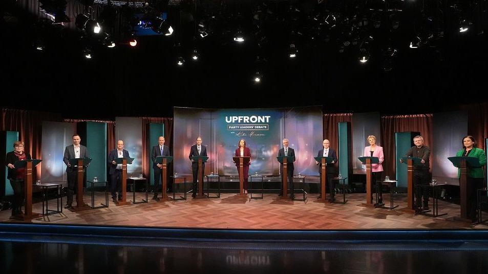 Ten party leaders stand at podiums during a televised  election debate. The floor and podiums are brown wood and the candidates are looking toward the camera