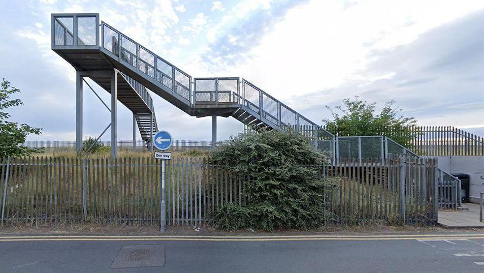 A railway bridge taken from a side profile. The silver metal structure has three flights of steps leading to the top which are above a grass bank. A large security fence stands in front of the bridge and there is also a blue one-way road sign in the foreground of the image.