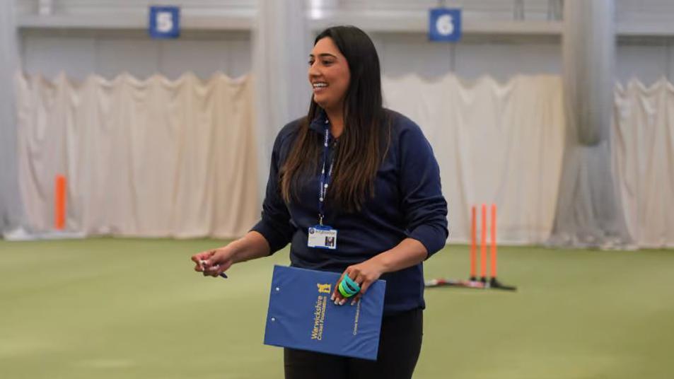 Simran is wearing a navy blue long-sleeved sports polo top and matching trousers. She has a navy blue lanyard around her neck and is holding a clipboard that has the yellow Warwickshire Cricket Foundation logo printed on the back of it. In the other hand she is holding a pen and she has a french manicure. She is smiling with her teeth and looking off to the side. Simran has long, dark brown hair. The background is out of focus, but there are white sheets and netting, as well as orange plastic cricket stumps on the green astro floor.