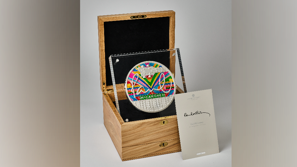 A silver coin which has Paul McCartney's signature on, along with a colourful striped background. It is presented in a wooden box.