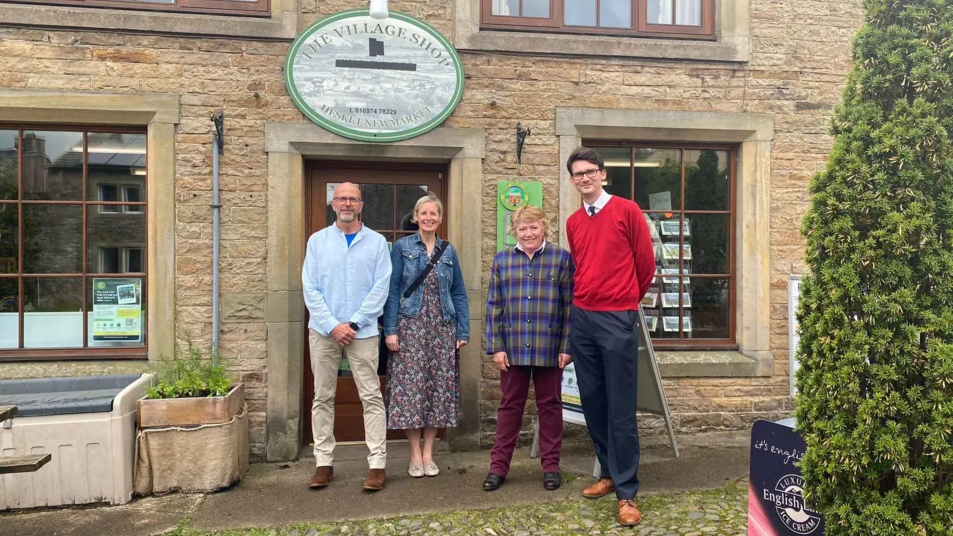 Four people, two men and two women, are stood outside a building with a sign saying 'The Village Shop' on it. 