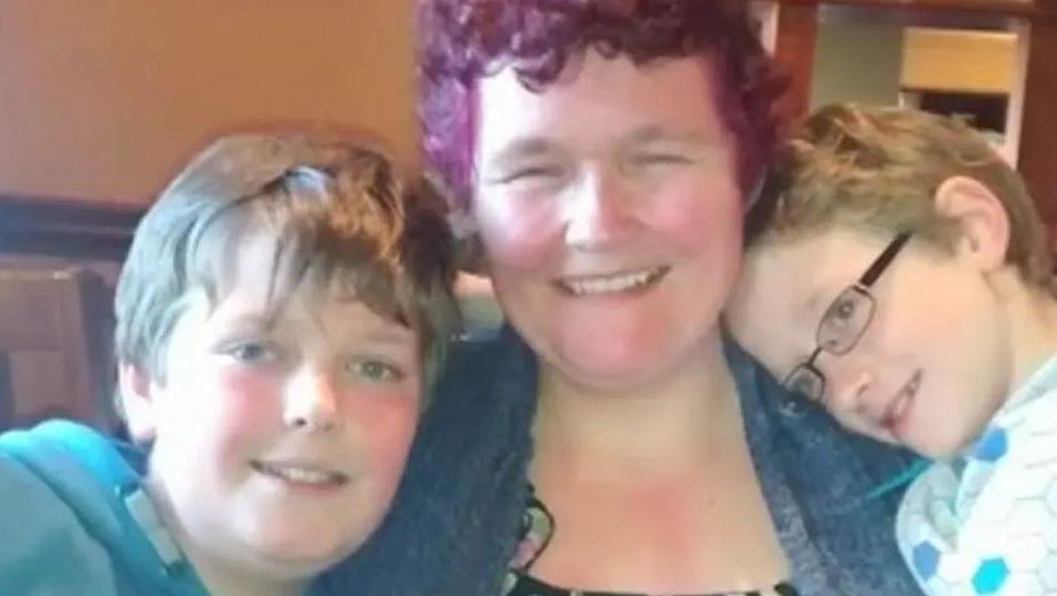 Claire Throssell smiles as she hugs her children Jack and Paul. She has short hair, died purple, and wears a blue denim shirt. Jack has brown hair, wears a blue top and is smiling into the camera. Paul has light-brown hair and wears glasses and a white and blue top.