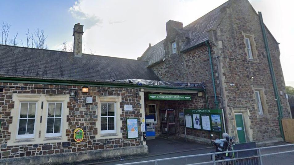 Barnstaple Station with a green and white sign above the entrance and a green and white information board. A defibrillator unit in yellow and green is attached to the wall and a woman is approaching the station on a mobility scooter.