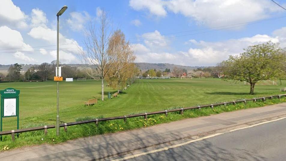 Master Park in Oxted, Surrey. On the left is a sign displaying events that are happening in the area, and on the right is a sizeable green space with benches and trees dotted through the middle. On the right side there is one big tree with green leaves, behind which football goals are visible.