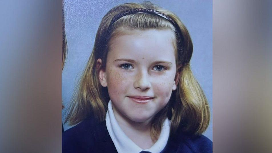 Ariane Murrell as a schoolgirl before she lost her hair. In this image, she is wearing a white shirt with a collar and blue jumper and is smiling at the camera. She has shoulder length light brown hair and is also wearing a hairband. 