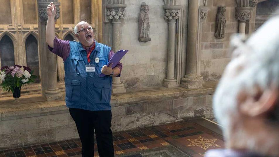 Stephen de Silva wears a maroon shirt and a blue gilet on which is attached gold coloured badge with the word guide on it. He also has a red lanyard round his neck attached to a plastic case containing his name badge and is holding a purple folder.  He has grey hair with a short grey moustache and beard and is balding. He is wearing clear-rimed glassed and is explaining something to visitors inside the cathedral.