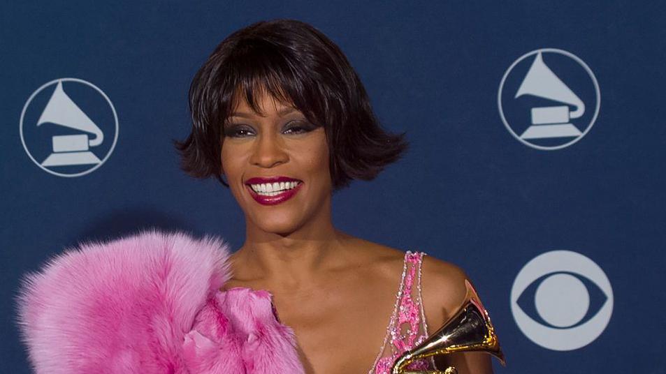 Whitney Houston backstage at the Grammy Awards Show in February 2000 in Los Angeles. She is holding her Grammy and wearing a pink fur thow over one shoulder. 