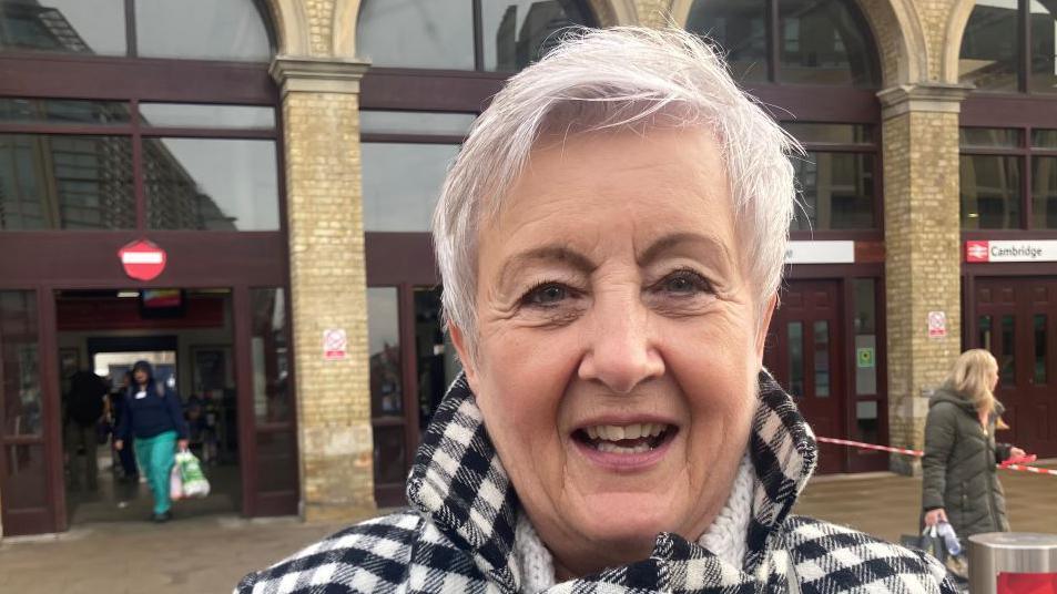 A woman stands outside Cambridge train station, she has cropped white hair and is wearing a black and white checked coat.