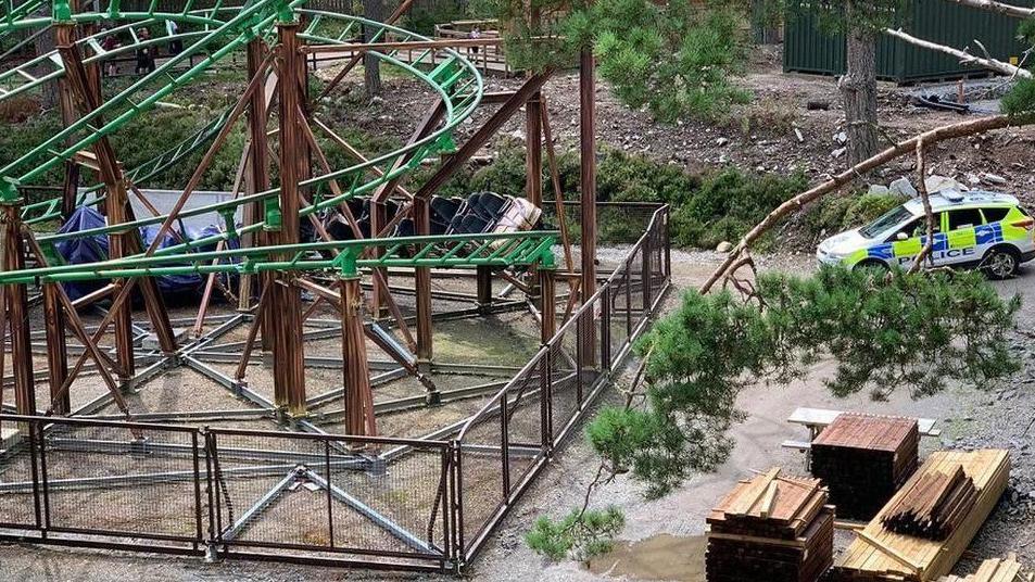 A police car is parked near to Landmark Forest Adventure Park's Runaway Timber Train rollercoaster. The ride is behind a high wire fence. The ride is a twisting, green track among pine trees.