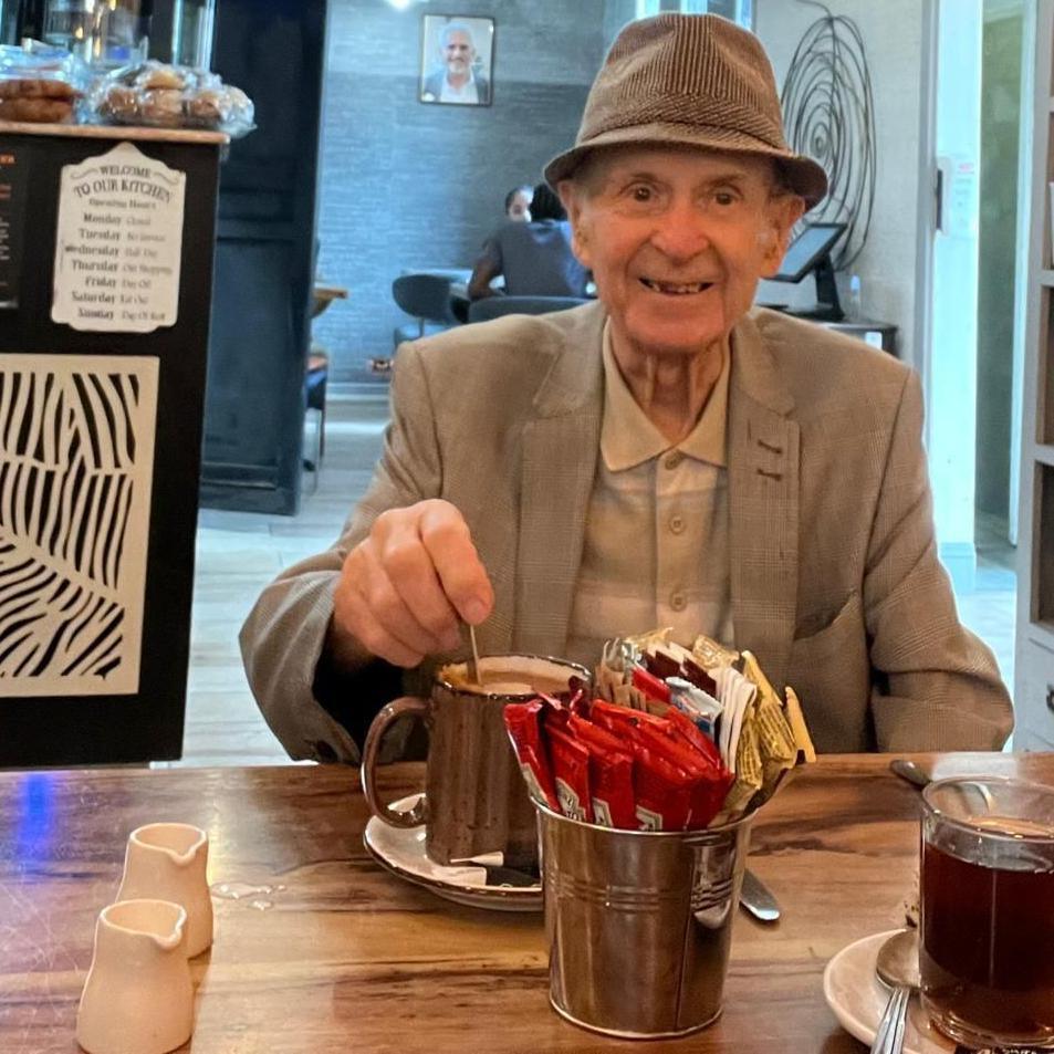 An elderly man smiles (right) for a photo in a cafe. He is wearing a pork-pie hat and a cream jacket and light shirt. He is stirring a hot drink in a mug on a table in front of him which contains a mental container of condiments