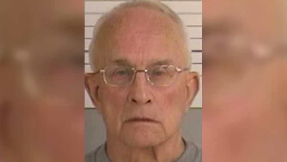 A custody image of an elderly man. He is looking at the camera. He has glasses, short grey hair and is wearing a grey tracksuit top.