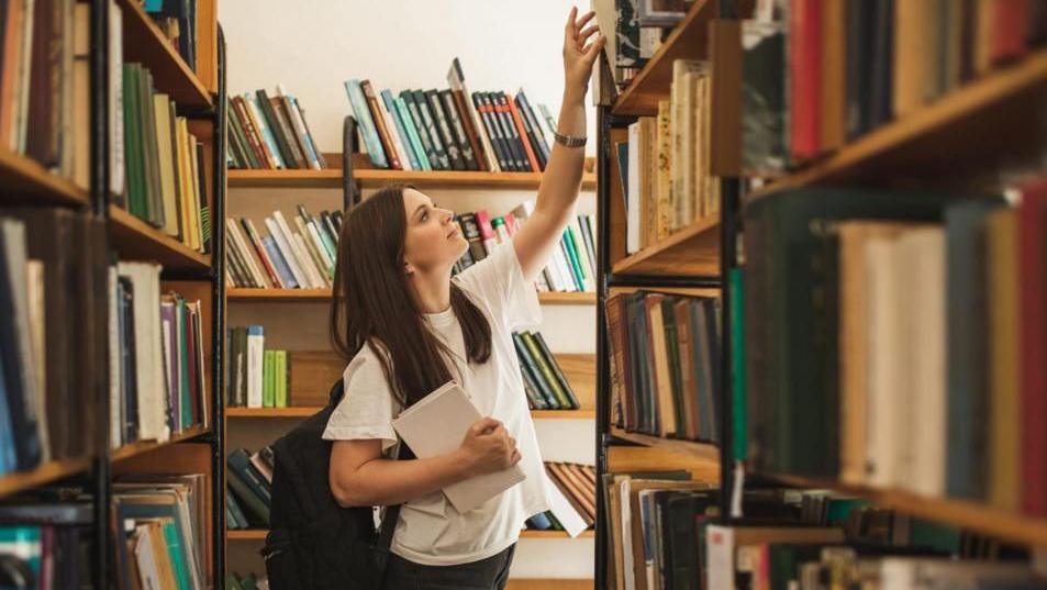 A woman with long brown hair, wearing a white t-shirt an dark jeans, with a black backpack on her shoulder is leaning up to take a book of a packed bookshelf. She is surrounded by other packed wooden bookshelves and is holding a hardback book with a white cover.