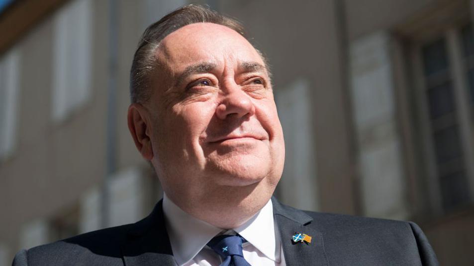 A close-up of Alex Salmond looking off into the distance, wearing a dark blue suit, white shirt and blue tie. 