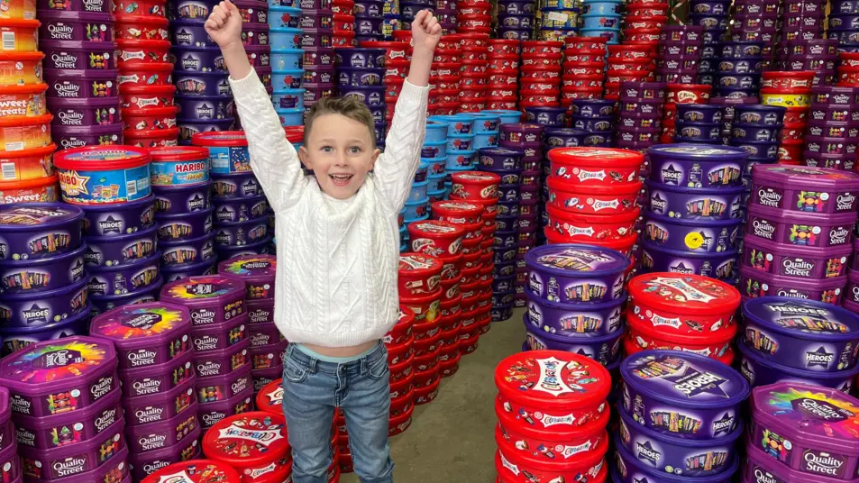 Teddy wears a white jumper and jeans. He has short light brown hair. His arms are raised in triumph. He is in a warehouse full of thousands of sweet tubs, including Heroes, Quality Street, and Celebrations.