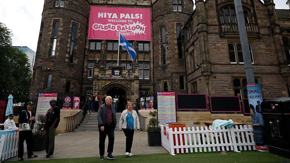 People milling about the front of an Edinburgh Fringe venue with a pink banner saying 'Hiya Pals' on the front of it. 