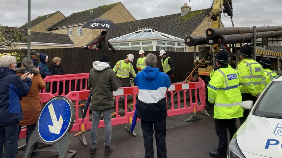 Workers, police and residents gather in a street in Broadway. There is a lorry carrying the poles and works are behind barricades.