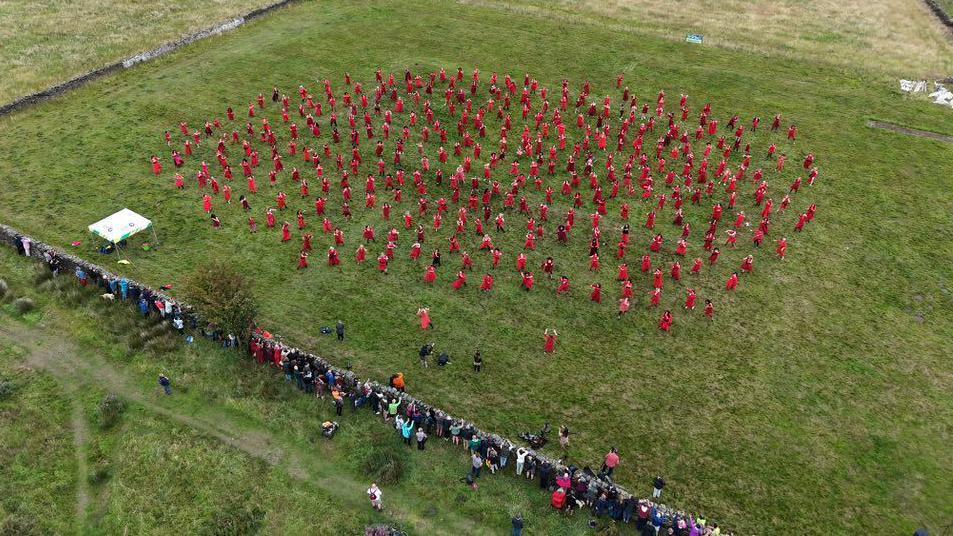 A drone photograph of a group of people dressed in red dressed dancing on the moors in Yorkshire. There is a line of people watching nearby.