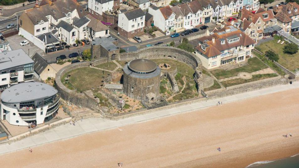 The circular Sandgate Castle, surrounded by a stone wall, located on a large sandy beach. The castle is seen from above and surrounded by other, more modern buildings.