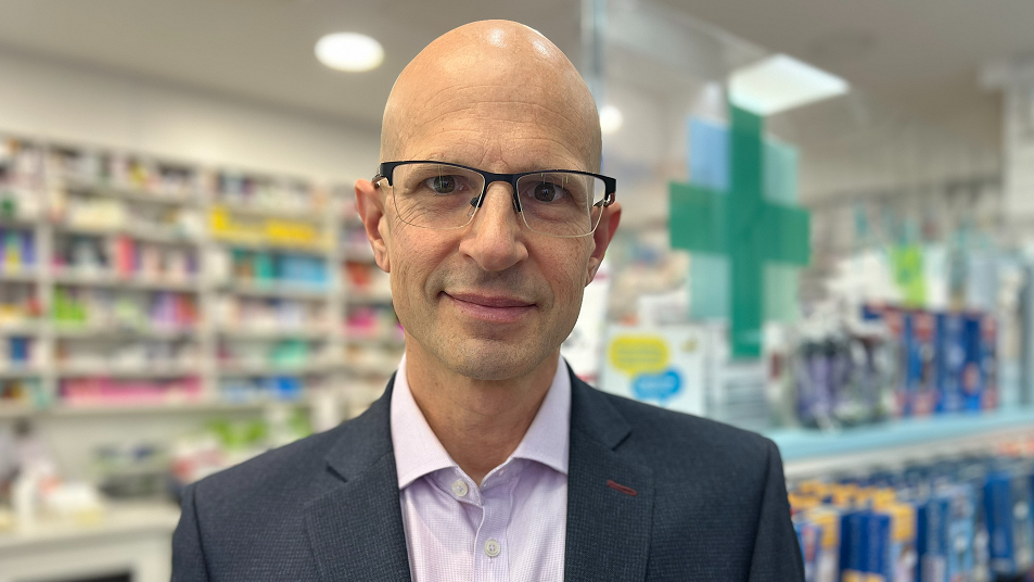 Olivier Picard is bald and is wearing a blue jacket and lilac shirt. He is wearing glasses and is standing in a pharmacy.