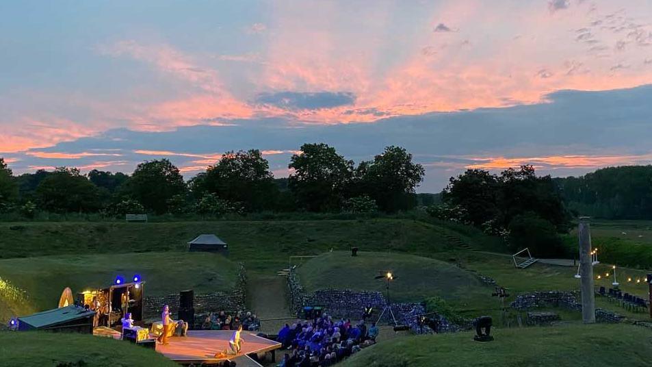A sunset can be seen over the Roman Theatre at Verulamium. The stage is set amongst the excavations and there are actors lit on the stage.