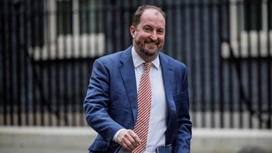 A smiling Guto Harri wearing a blue suit and striped tie walking down Downing Street