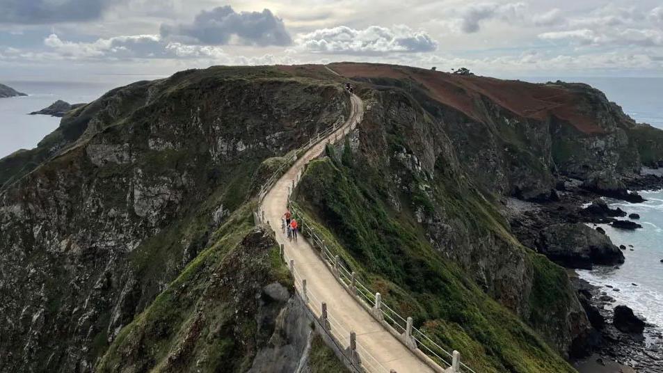 A high up view of the road between Sark and Little Sark, a long light brown stone path surrounded by descending rocks which are surrounded by the ocean.