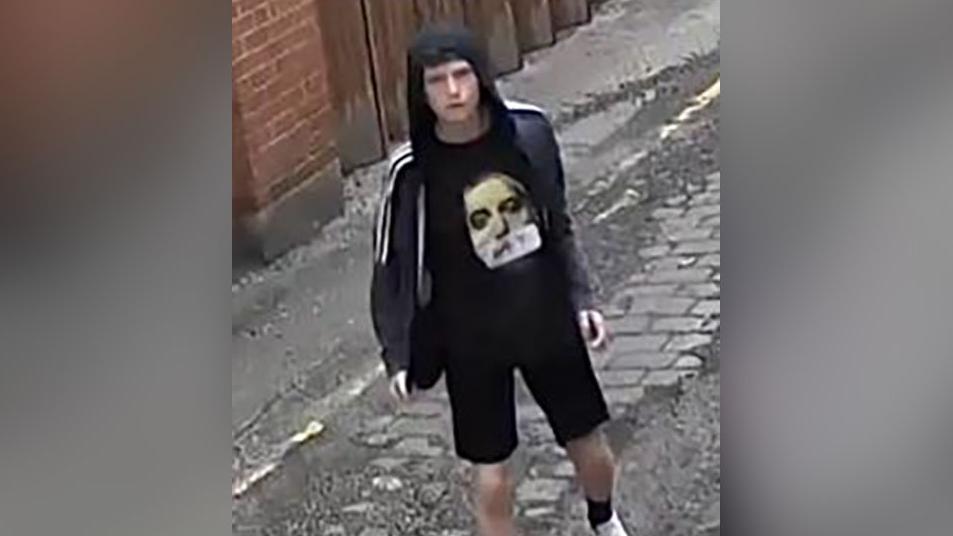 CCTV image of a man with black hair wearing an unzipped, hooded black jacket with white stripes running halfway down each sleeve. He is also wearing a black t-shirt with a man's grey face on it, and black socks and white trainers. He is walking down an alley. 