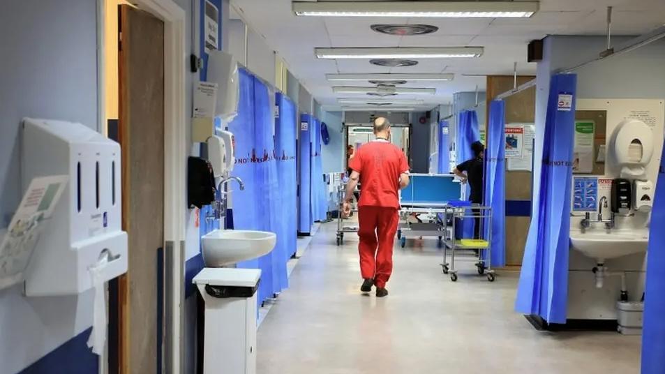 A male doctor in red hospital scrubs walks away from the camera as he heads down a hospital A&E ward with curtained rooms either side