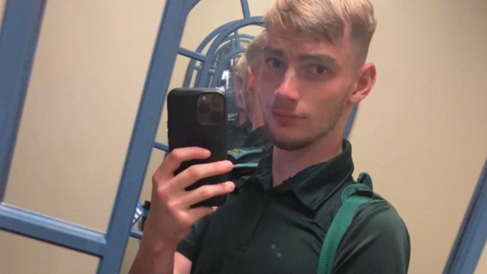 Jamie Kadolski takes a selfie in a mirror. He has short blond hair and is wearing a green polo neck with green epaulettes on either shoulder. His phone is black.  