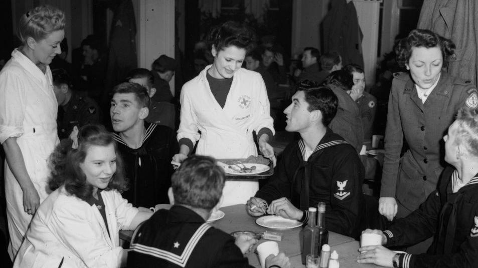 American Red Cross waitresses at the Rainbow Corner, the American Servicemen's Club in London, taking customers' orders in early 1945. Four US sailors are seated at a table. There are three waitresses, possibly British, in white, two are standing and one is seated. They each talking to a sailor. A fourth woman in American Red Cross uniform is on the right talking to the fourth sailor. Behind them can be seen other seated US service personnel.