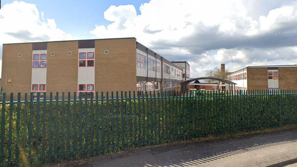 The school building seen from the side, behind a long green metal fence. There are two blocks from brown brick with long lines of windows and a green space between them. 