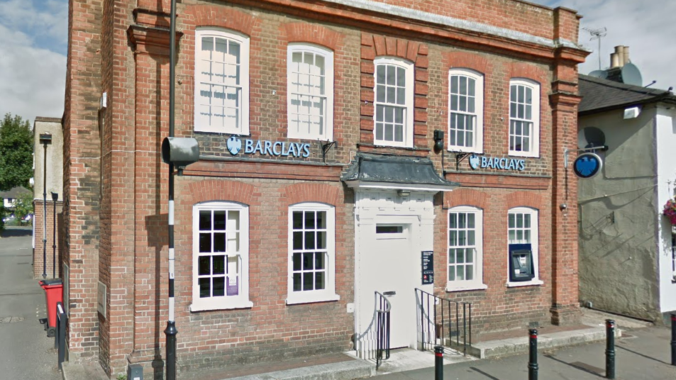 A branch of Barclays bank which is a two storey building with a white door and white framed windows with a cashpoint outside.