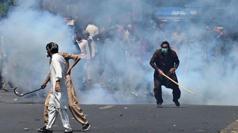 Pakistan Tehreek-e-Insaf party activists and supporters of former Pakistan's Prime Minister Imran hold rods and stand amid teargas fired by police during a protest against Khan's arrest in Lahore on 9 May 2023.