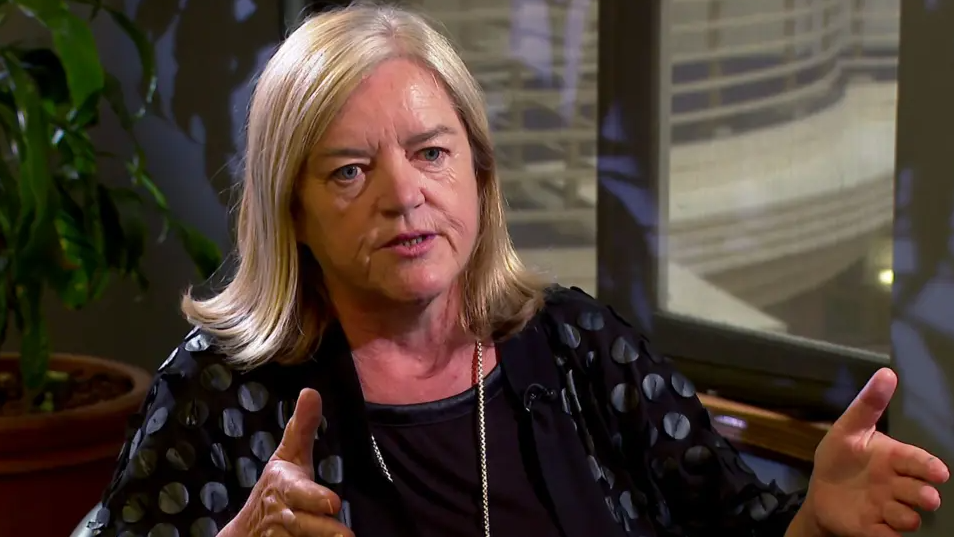 Ms Casey is sat in a chair being interviewed and is making a gesture as she speaks. She has shoulder length blonde/brown hair and is wearing a dark coloured top.