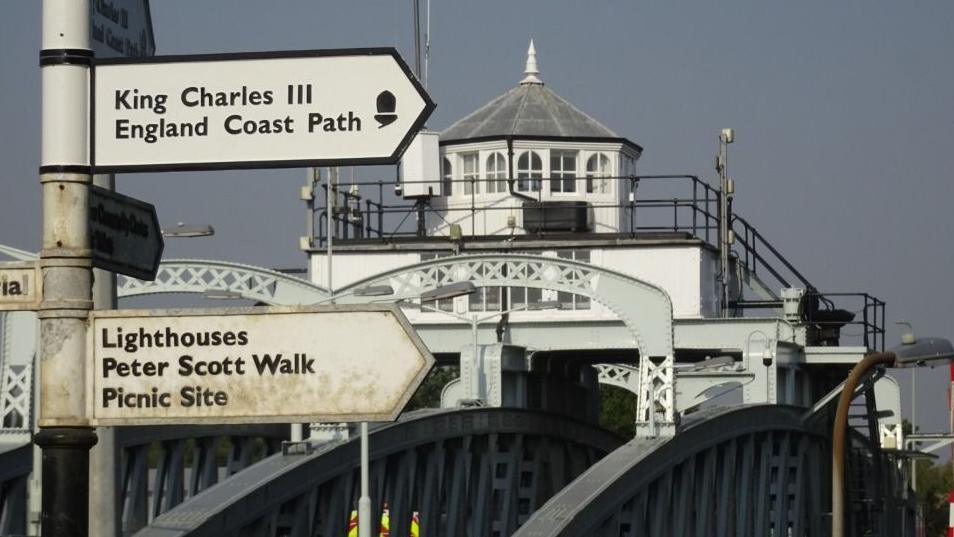 Signposts indicating the way to the King Charles III England Coast Path and other attractions at Sutton Bridge. In the background is the Cross Keys Bridge, a white and grey metal swing bridge with a Victorian engine house on top. 