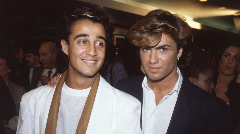 Two men with brown hair, the man on the left is smiling and looking to the side. He's wearing a white suit and a white t-shirt. The other man is wearing a black suit and a white shirt - he's looking serious and straight at the camera.