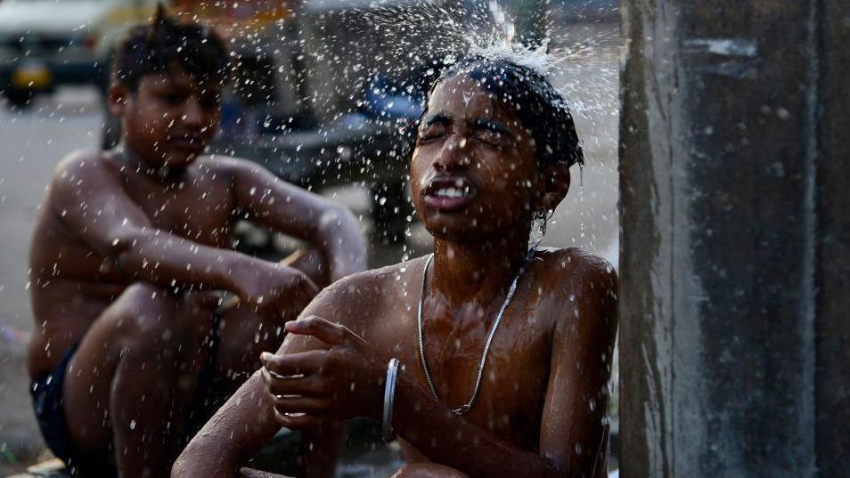 Two shirtless boys cool off under a water tap in India, the boy in the foreground has his eyes shut as a torrent of water splashes onto his head.