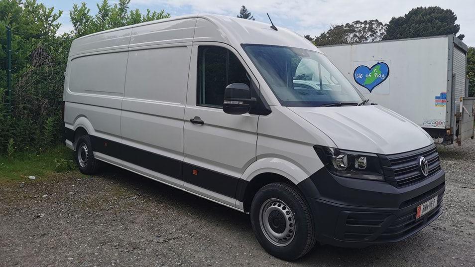 The side of a white Peugeot boxer van parked on gravel in front of a white trailer with the Isle of Play logo on it. The logo is a blue heart shape with the shape of the Isle of Man in the middle and the words Isle of Man written across the middle in white lettering.