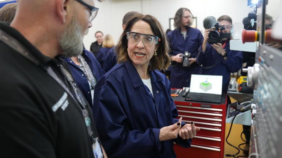 Liz Kendall at Peterborough College discussing the work being done there. She is wearing protective overalls and glasses and in the background a number of people are watching, including someone with a television camera.