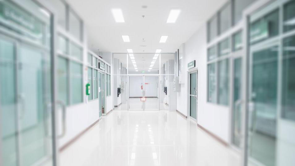 Image shows clear doors opened on the left and right of the picture showing a hospital corridor with a white floor, walls and ceiling with glass doors on the left and right 